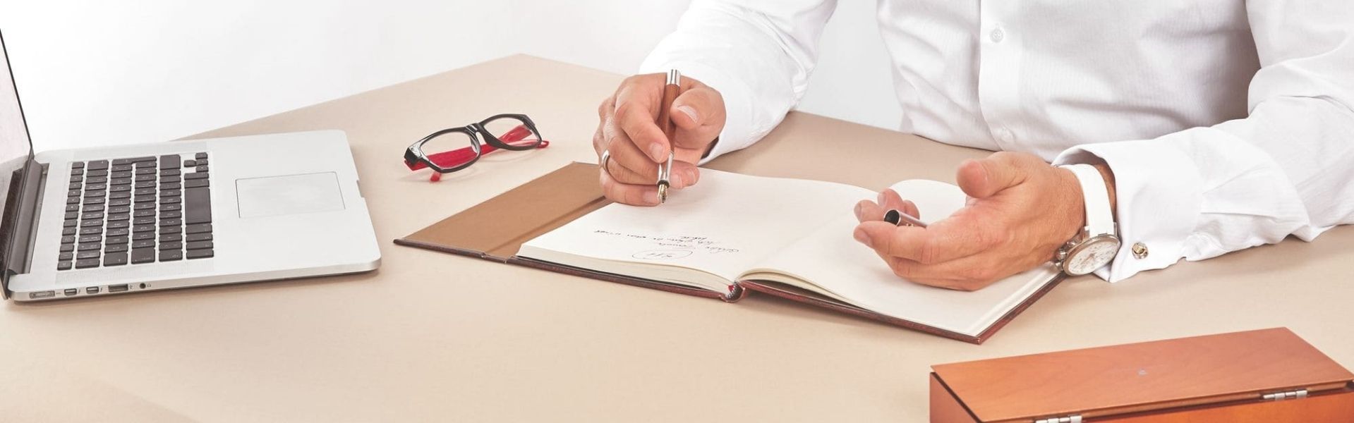 Schreibtisch mit aufgeklapptem Laptop, Brille, geöffnetem Buch und zwei Händen über dem Buch